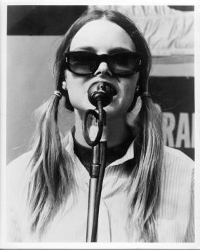 michelle_phillips_singing_at_a_microphone_wearing_sunglasses_and_hair_in_pigtails